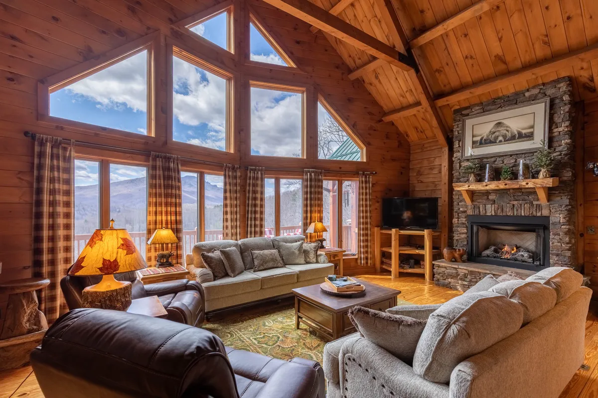 A cozy living area with large windows framing mountain views, a central stone fireplace, and comfortable sofas and armchairs arranged for conversation. A decorative lamp adds warmth and ambiance, while the fireplace anchors the room as a gathering point.