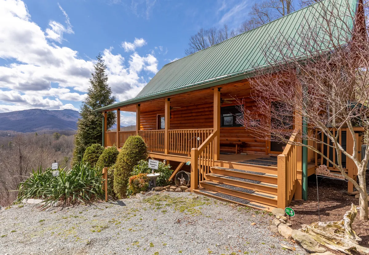 A welcoming exterior view of the log cabin structure with a green metal roof and a landscaped garden. Wooden steps and a porch invite guests to approach and relax, emphasizing the home’s inviting and accessible nature for all family members.