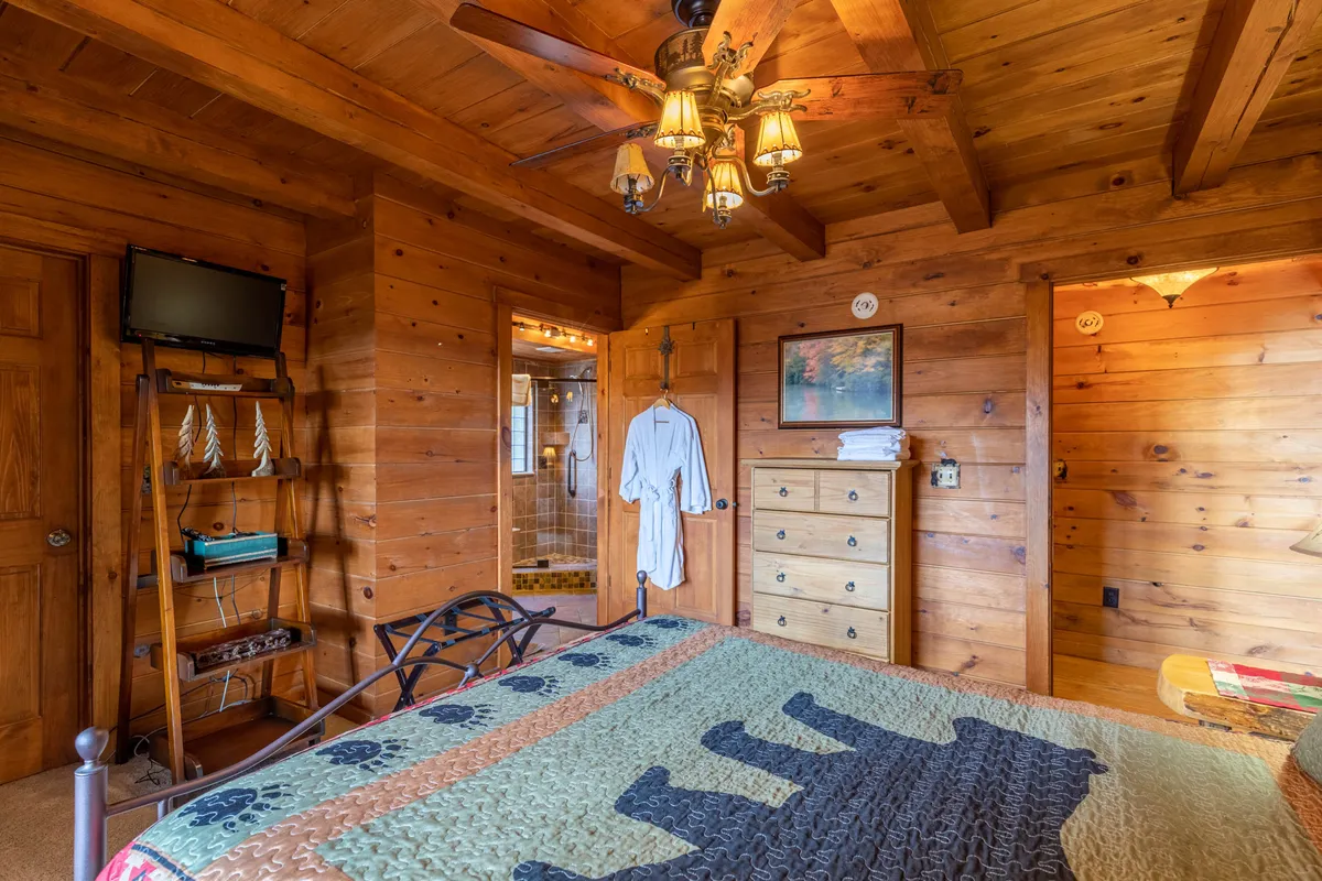A cozy bedroom with wooden ceiling beams, a bed dressed in a patterned quilt, a wooden dresser, and a charming ceiling fan. The room exudes a warm, homey feel, perfect for relaxation after outdoor adventures.