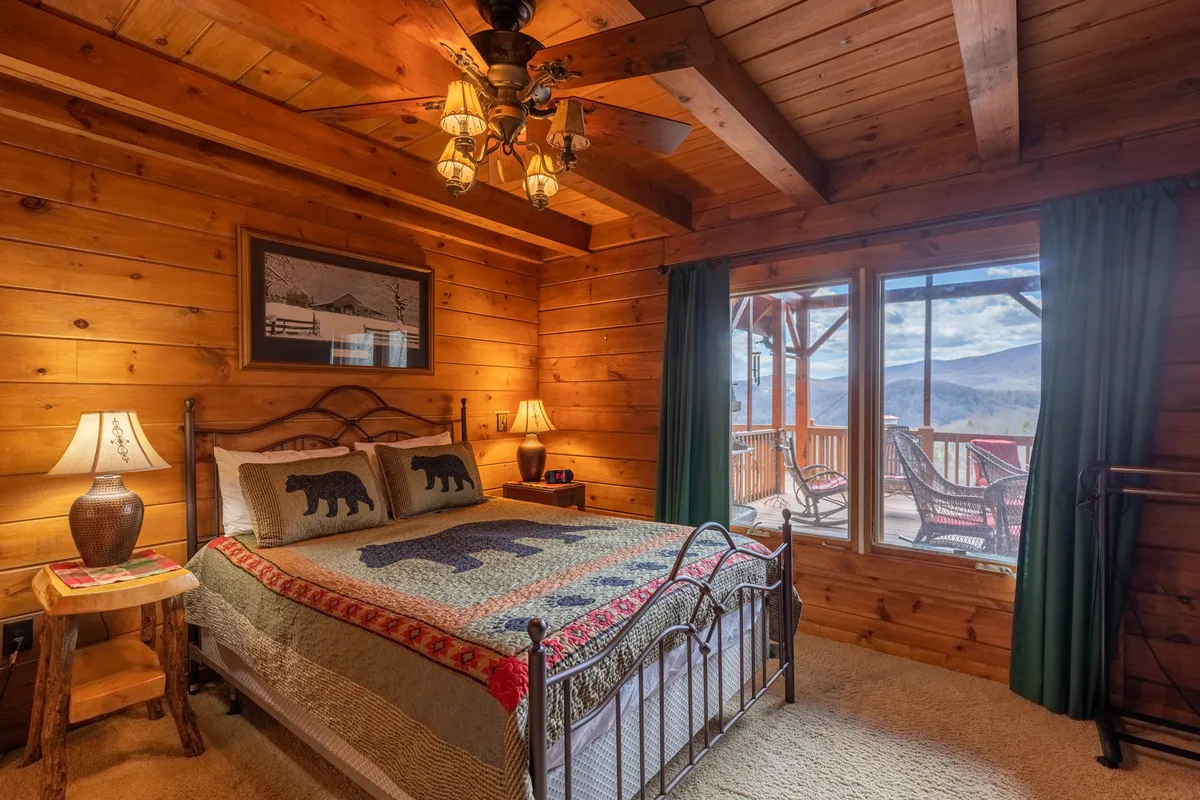 A warmly lit bedroom featuring a bed with a bear-themed quilt, large window offering mountain views, rustic wooden beams, and bedside tables with lamps. The decor is inviting and has a touch of mountain lodge charm, ideal for restful nights.