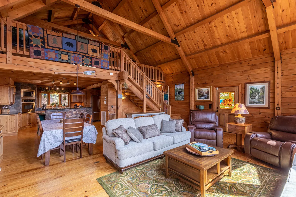 A wide shot of the open-concept cabin living space, featuring a large dining table for six, plush sofa, wooden ceiling beams, and a staircase leading to an upper level. The space is illuminated by natural light streaming through windows, with the dining, living, and kitchen areas all visible and seamlessly connected.