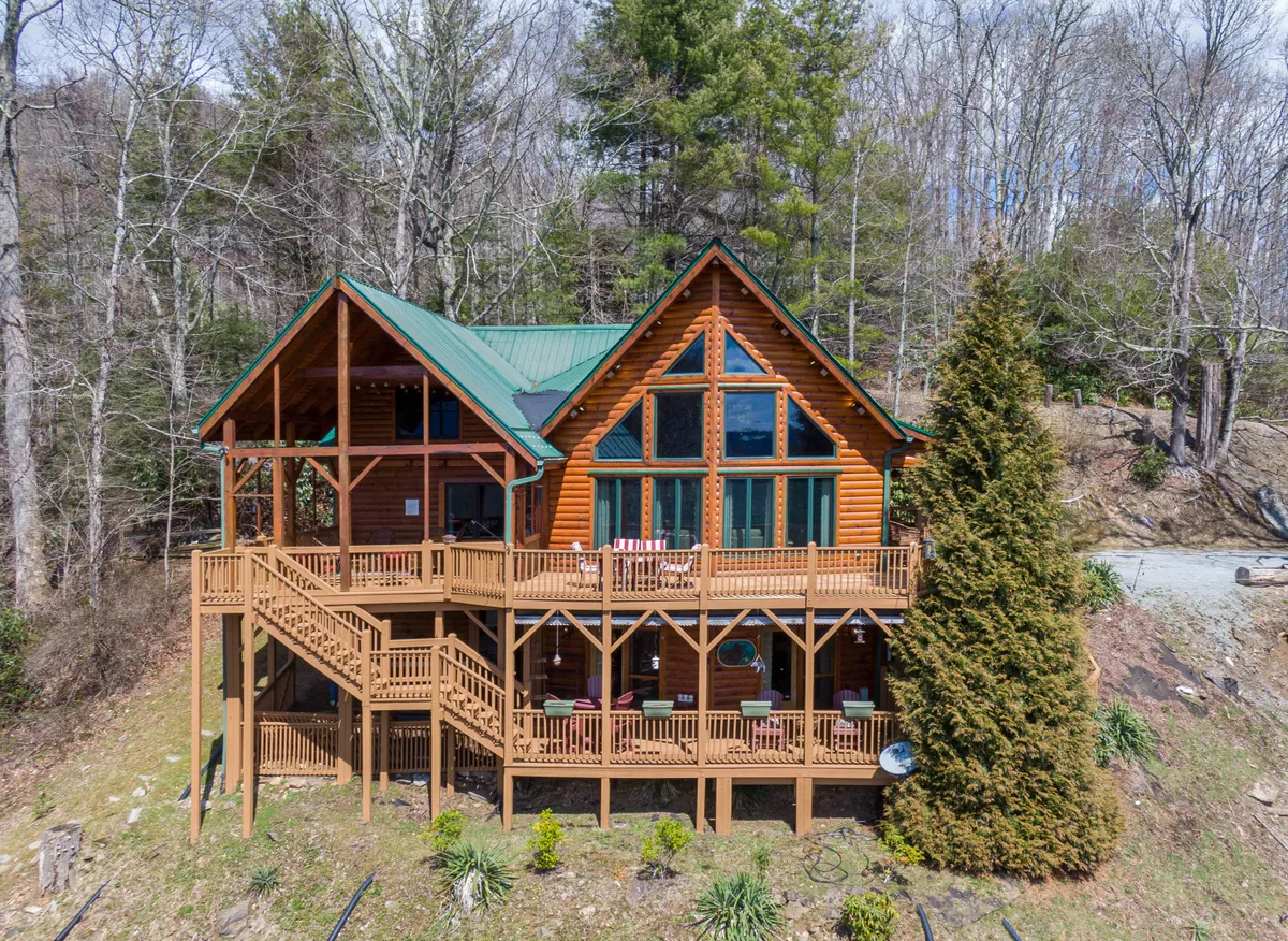 The image showcases the inviting log cabin exterior, featuring classic rustic architecture, a striking green metal roof, expansive windows, and a spacious wooden deck surrounded by towering trees. The deck overlooks mountain vistas, promising breathtaking sunset views and tranquil mornings immersed in nature.