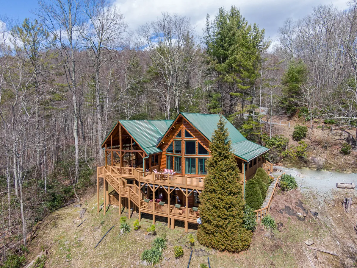 This image displays the welcoming log cabin structure of Beary Relaxing Cabin with its green metal roof, surrounded by trees and featuring a spacious deck with comfortable seating. The outdoor area and the natural setting dominate the view, hinting at forest trails and the proximity to mountain adventures.