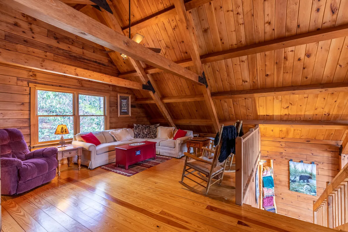 A cozy living area featuring a sofa and rocking chair set beneath exposed wooden beams. Expansive windows provide sweeping views of the surrounding landscape, inviting the outdoors in and offering a tranquil spot to enjoy nature from the comfort of the cabin.