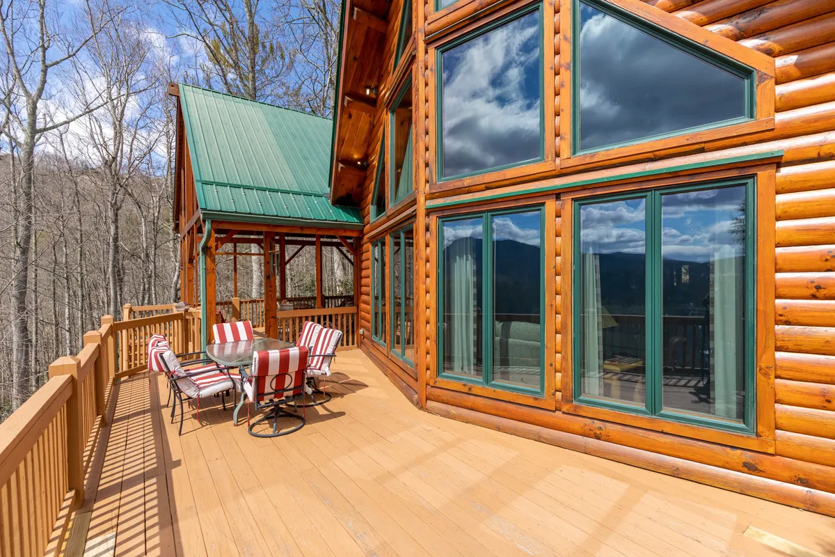 This exterior image features the Beary Relaxing Cabin's large glass windows, wooden log exterior, green metal roof, and an inviting outdoor seating area, all set against a backdrop ideal for enjoying mountain views. The scene also hints at activities like outdoor dining and relaxing while immersed in nature.