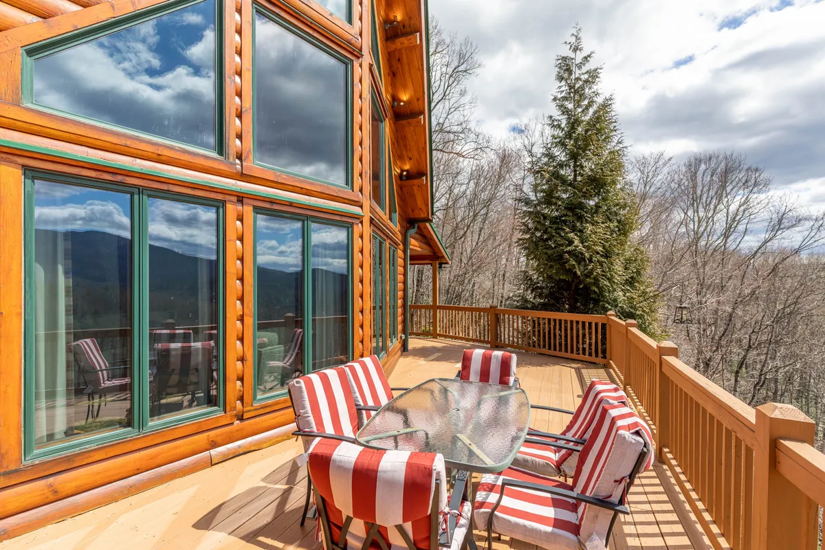 This image captures the cabin's exterior deck featuring large windows, striped outdoor chairs, and a sturdy deck railing. The outdoor space is open and inviting, surrounded by mountain views and lush greenery, offering plenty of room for relaxation and outdoor activities.