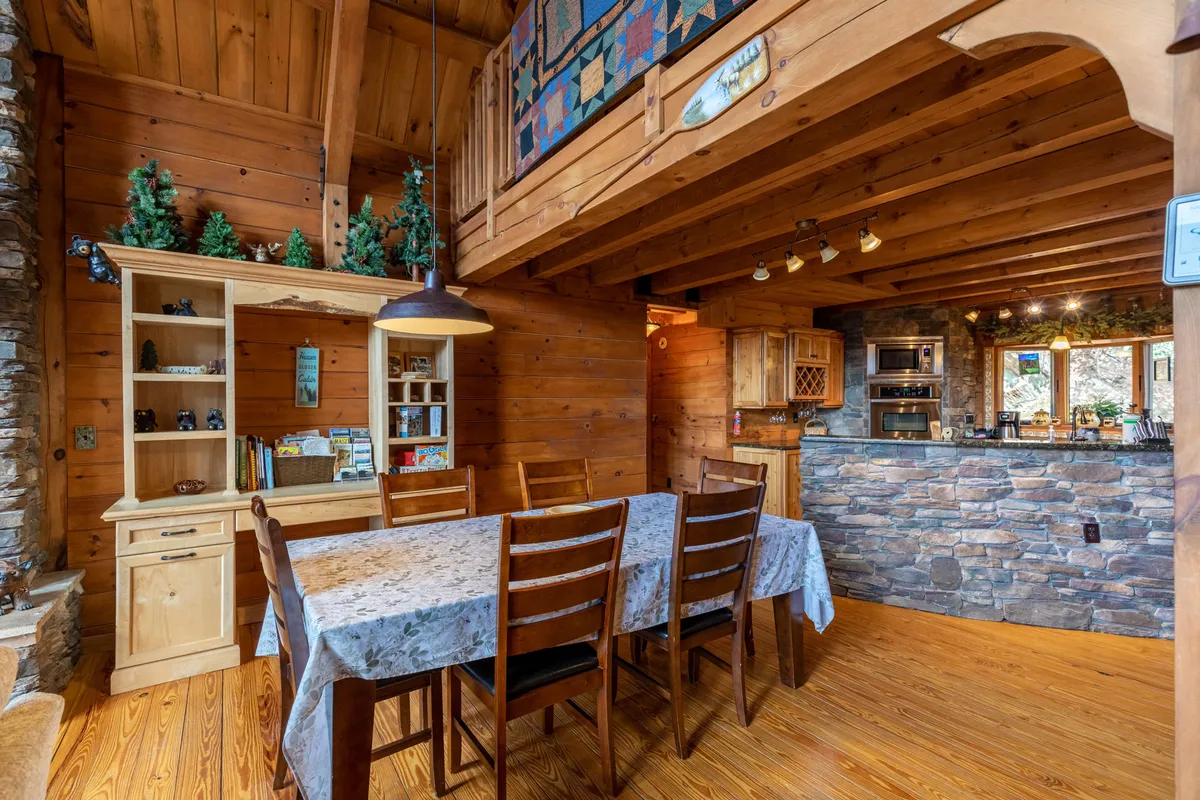 A view of the dining table set in a warm, wood-accented space, adjacent to an open kitchen and beneath exposed wooden beams. The area is designed for both meals and socializing, with the open layout inviting easy movement between spaces.