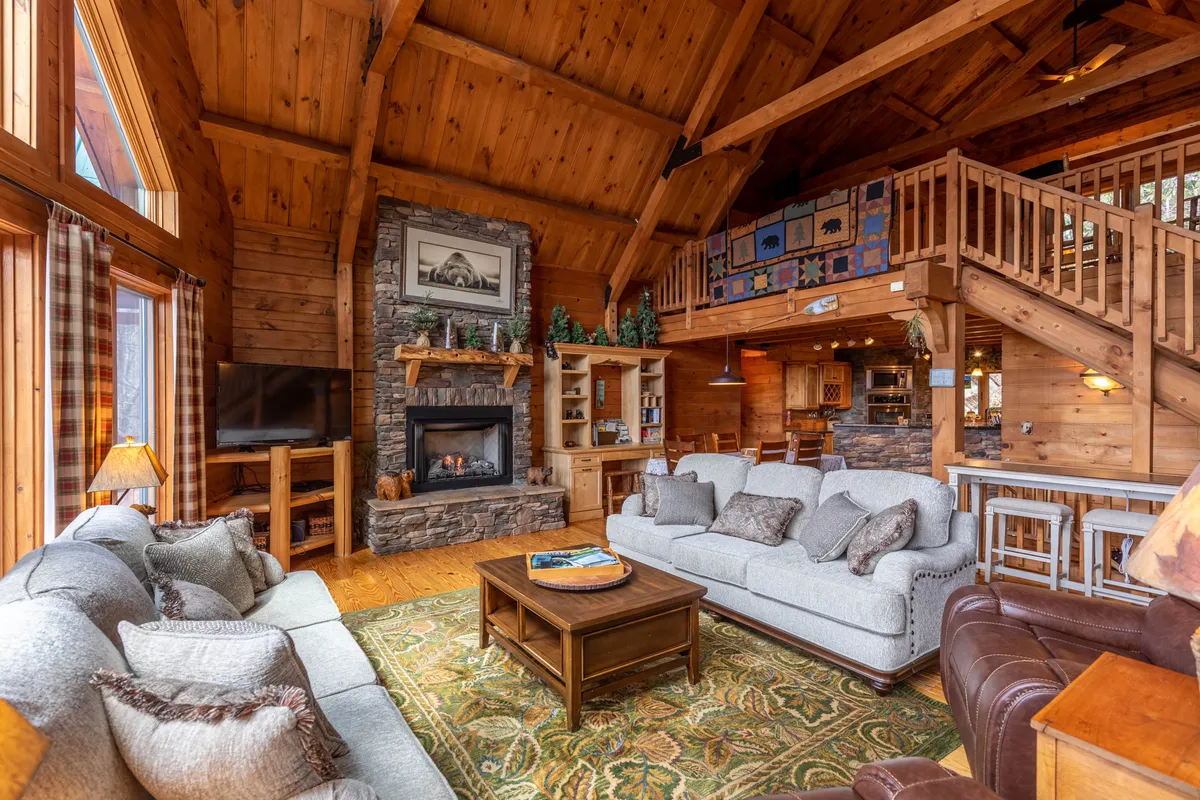 A warm, welcoming living room with a striking stone fireplace, large windows framing mountain scenery, plush seating, and exposed wooden beams. The setting is perfect for relaxing after a day of adventure, hosting family gatherings, or cozying up by the fire in winter.