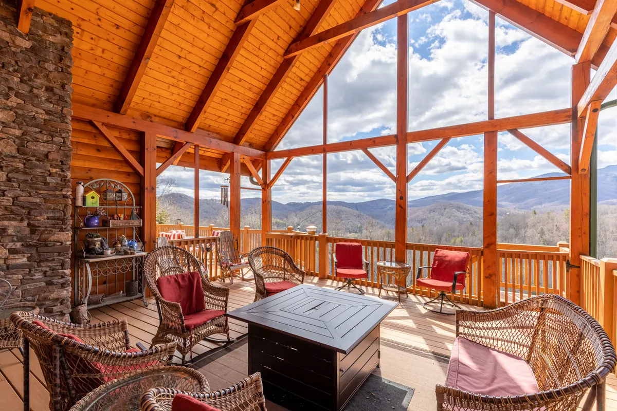 A sweeping mountain vista is seen from the cabin’s deck, with expansive windows, rustic wooden beams, and a cozy seating arrangement showcasing the stunning views available from the property. Decorative shelves add a homely touch, while the scene connects indoor comfort with the rugged outdoors.